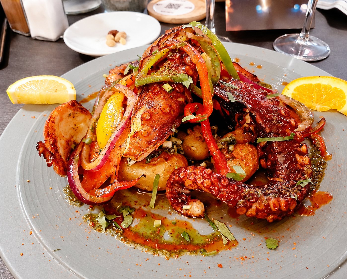 Un poulpe grillé servi sur une assiette grise avec des poivrons, des oignons rouges, des pommes de terre, des herbes fraîches et des quartiers de citron et d’orange.
