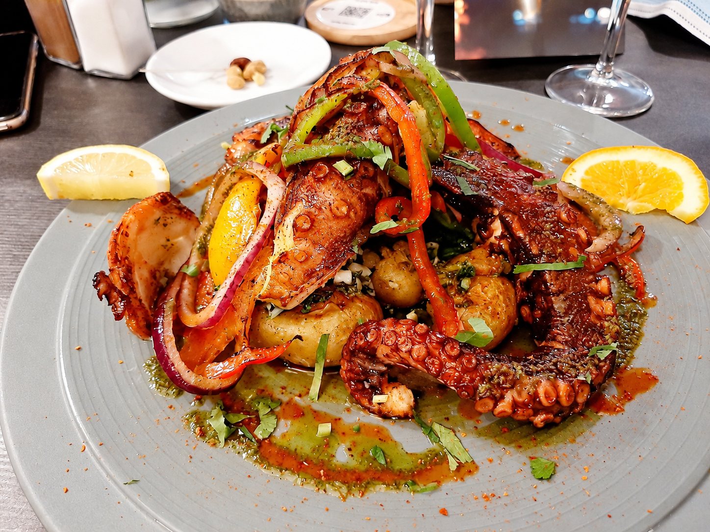 Une assiette grise garnie de tentacules de poulpe grillés, de lanières de poivrons, d’oignons rouges et de pommes de terre, accompagnée de quartiers de citron et d’orange sur une table de restaurant.