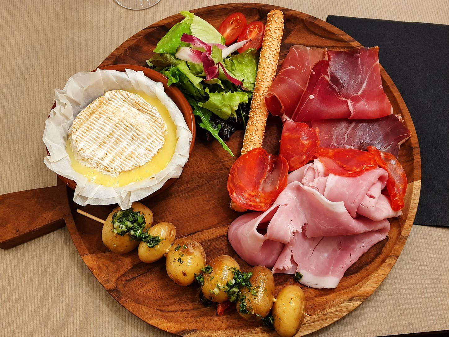 Vue de dessus d’une planche en bois ronde avec un camembert fondu, de la charcuterie, une brochette de pommes de terre grenaille, une salade et un gressin.