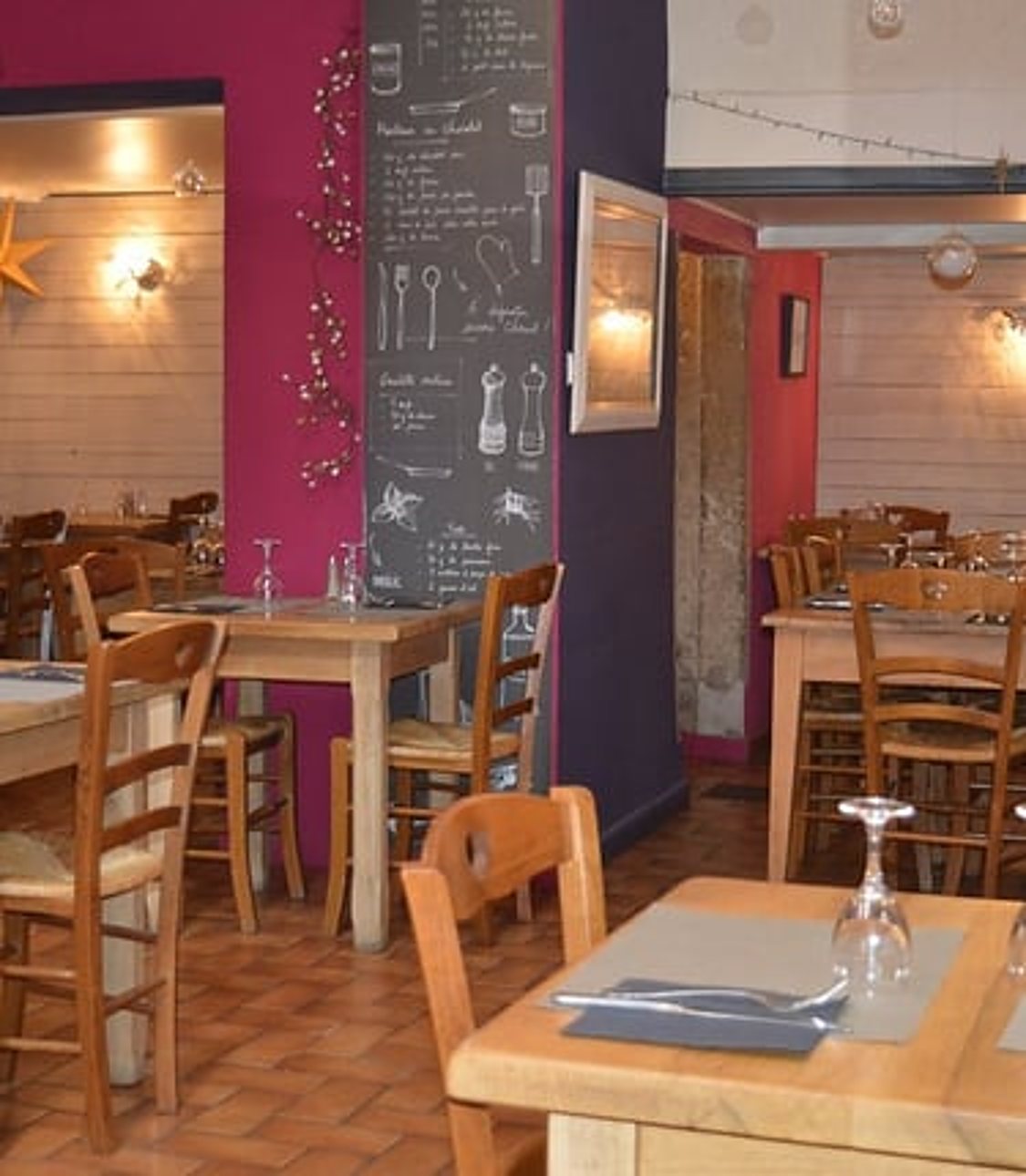 Intérieur d’un restaurant avec des tables et chaises en bois dressées, un mur framboise et une grande ardoise décorée de dessins à la craie.