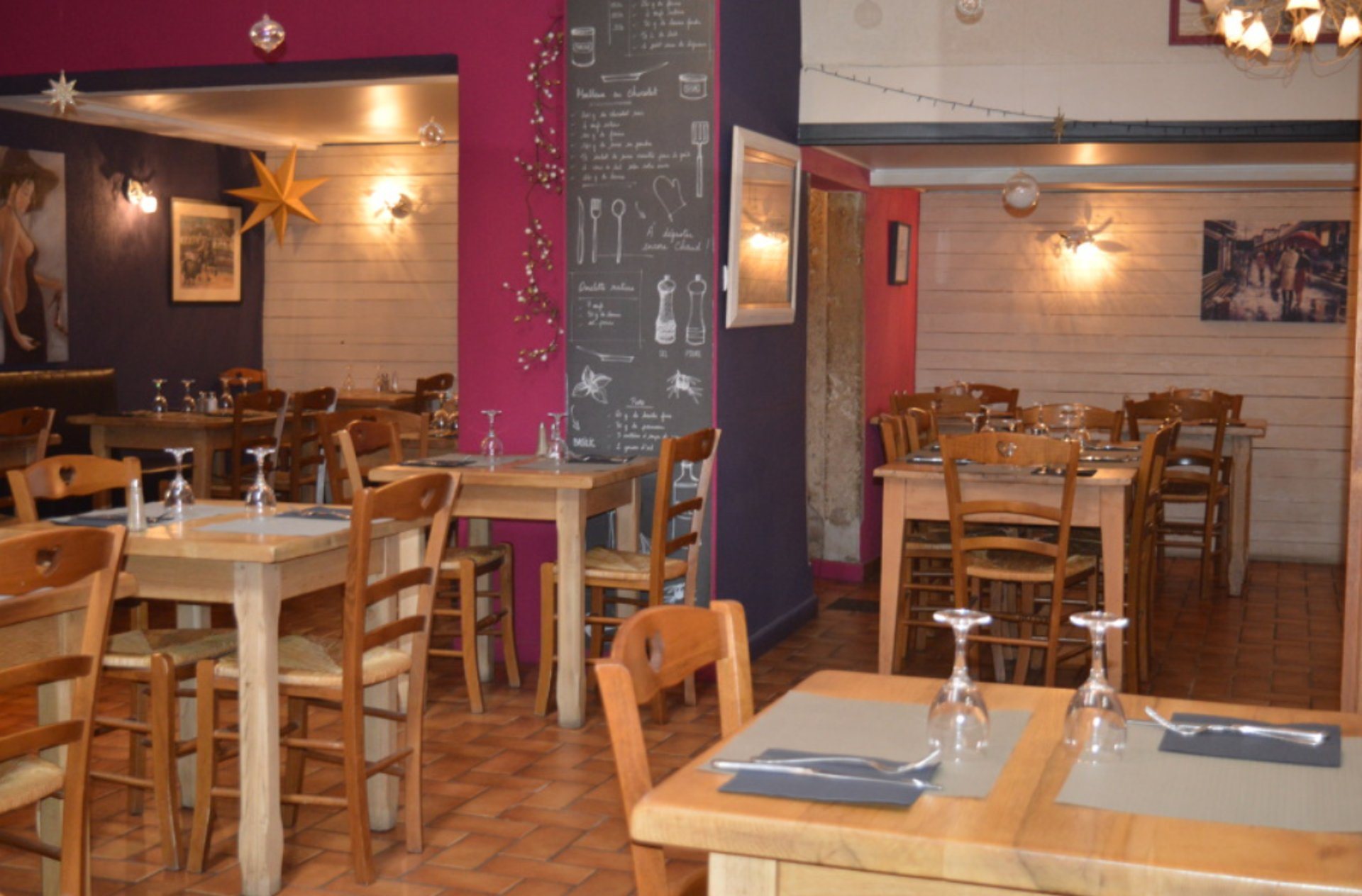 Salle de restaurant aux murs framboise et lambris clairs, avec tables et chaises en bois dressées et une grande ardoise de recettes au mur.