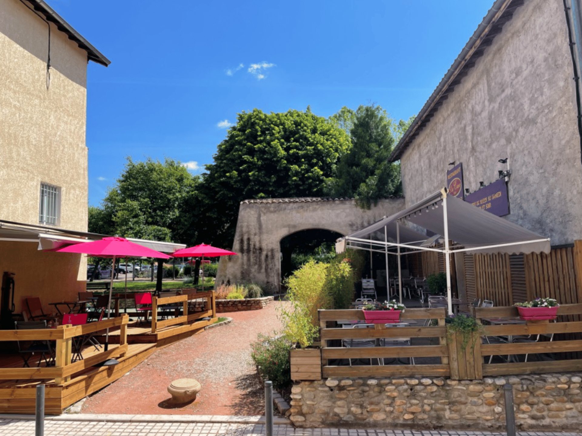 Terrasse de restaurant en plein soleil avec parasols roses, clôtures en bois et jardinières fleuries, devant une arche en pierre et des arbres.